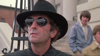 Movie still from “Wise Blood” (1979), directed by John Huston – A man wearing sunglasses and a hat is standing in front of some steps; Close Up shot, Low angle