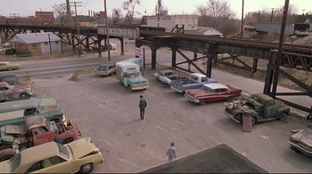 Movie still from “Wise Blood” (1979), directed by John Huston – A man walking across a street next to cars and a bridge; Extreme Wide shot, High angle