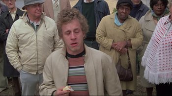 Movie still from “Wise Blood” (1979), directed by John Huston – A group of people standing around a man eating a sandwich; Close Up shot, High angle