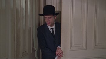 Movie still from “Wise Blood” (1979), directed by John Huston – A man in a suit and a hat leaning against a wall; Medium shot, Low angle