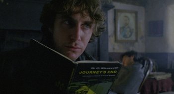 Movie still from “Withnail & I” (1987), directed by Bruce Robinson – A man reading a book in a room with other people in the background; Close Up shot, Over the shoulder angle