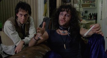 Movie still from “Withnail & I” (1987), directed by Bruce Robinson – A man sitting on top of a brown couch next to another man; Medium shot, Over the shoulder angle