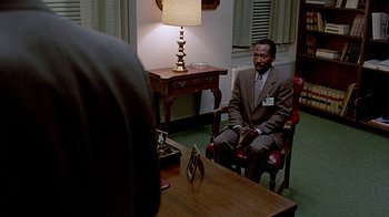 Movie still from “Witness” (1985), directed by Peter Weir – A man sitting in a chair in front of a desk; Medium shot, Over the shoulder angle