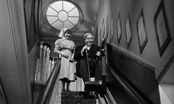 Movie still from “Witness for the Prosecution” (1957), directed by Billy Wilder – An older man sitting on top of an escalator next to an older woman; Wide shot, Low angle