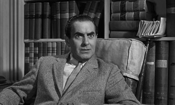 Movie still from “Witness for the Prosecution” (1957), directed by Billy Wilder – A black and white photo of a man sitting in a chair in front of a book shelf; Medium shot, High angle
