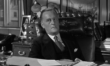 Movie still from “Witness for the Prosecution” (1957), directed by Billy Wilder – An older man sitting at a desk in front of a chair; Medium shot, Low angle