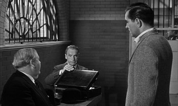 Movie still from “Witness for the Prosecution” (1957), directed by Billy Wilder – A man in a suit and tie holding a briefcase in front of two other men; Medium shot, Over the shoulder angle