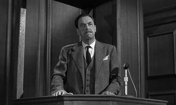 Movie still from “Witness for the Prosecution” (1957), directed by Billy Wilder – A man in a suit and tie standing at a podium with a microphone; Medium shot, Low angle