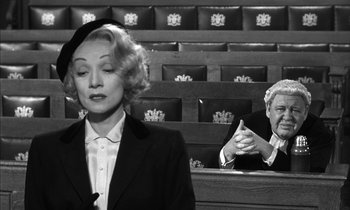 Movie still from “Witness for the Prosecution” (1957), directed by Billy Wilder – A woman in a courtroom with a judge in the background; Medium shot, Over the shoulder angle