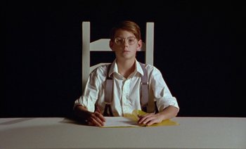 Movie still from “Wittgenstein” (1993), directed by Derek Jarman – A man sitting at a table in front of a piece of paper; Medium shot, Low angle