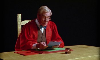 Movie still from “Wittgenstein” (1993), directed by Derek Jarman – An old man sitting at a table writing on a piece of paper; Medium shot, Low angle