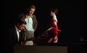 Movie still from “Wittgenstein” (1993), directed by Derek Jarman – Three men and a woman are standing in front of a piano; Medium shot, Low angle