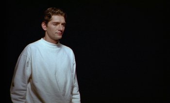 Movie still from “Wittgenstein” (1993), directed by Derek Jarman – A man in a white shirt standing in front of a black background; Medium shot, Low angle