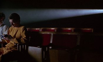 Movie still from “Wittgenstein” (1993), directed by Derek Jarman – A person sitting in a theater with a red seat; Wide shot, Low angle