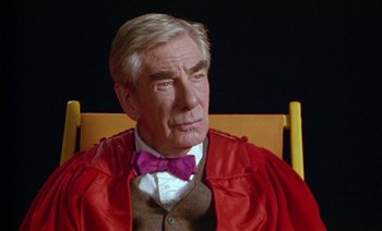 Movie still from “Wittgenstein” (1993), directed by Derek Jarman – An older man sitting in a chair wearing a red robe and a purple bow tie; Close Up shot, High angle