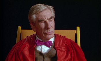 Movie still from “Wittgenstein” (1993), directed by Derek Jarman – An older man sitting in a chair wearing a red robe and a purple bow tie; Close Up shot, Over the shoulder angle