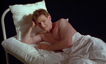 Movie still from “Wittgenstein” (1993), directed by Derek Jarman – A man laying in a bed with his hand on his head; Medium shot, High angle