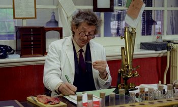 Movie still from “Wonderwall” (1968), directed by Joe Massot – A man in a lab coat writing on a piece of paper in front of a microscope; Medium shot, Low angle