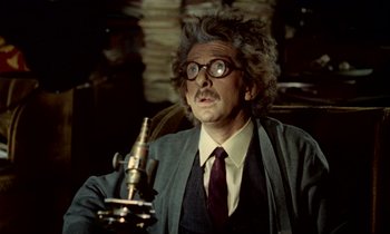 Movie still from “Wonderwall” (1968), directed by Joe Massot – A man in a suit and tie holding a microscope; Close Up shot, High angle
