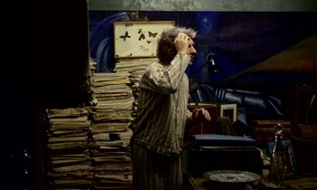 Movie still from “Wonderwall” (1968), directed by Joe Massot – An older man standing in front of a stack of papers; Medium shot, Overhead angle