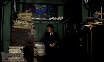 Movie still from “Wonderwall” (1968), directed by Joe Massot – A man in a suit and hat sitting in front of a pile of papers; Wide shot, High angle