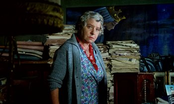 Movie still from “Wonderwall” (1968), directed by Joe Massot – An older woman standing in front of a pile of papers; Medium shot, Low angle