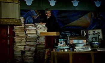 Movie still from “Wonderwall” (1968), directed by Joe Massot – A man standing in front of a stack of papers; Wide shot, Low angle