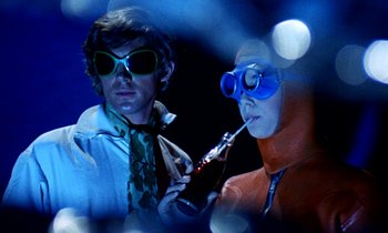 Movie still from “Wonderwall” (1968), directed by Joe Massot – A man and a woman dressed in costume drinking soda; Medium shot, Low angle