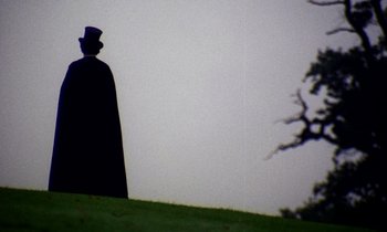 Movie still from “Wonderwall” (1968), directed by Joe Massot – A man wearing a top hat and a long black cape standing on top of a hill; Medium shot, Low angle