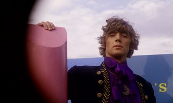 Movie still from “Wonderwall” (1968), directed by Joe Massot – A man in a purple shirt and a black jacket holding a pink object; Medium shot, Low angle