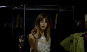 Movie still from “Wonderwall” (1968), directed by Joe Massot – A woman holding an apple while standing in a glass case; Medium shot, Low angle