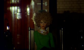 Movie still from “Wonderwall” (1968), directed by Joe Massot – A woman with a green shirt and a necklace is sitting in front of a wall; Medium shot, Low angle