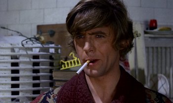 Movie still from “Wonderwall” (1968), directed by Joe Massot – A young man smoking a cigarette while wearing a maroon shirt; Close Up shot, Low angle
