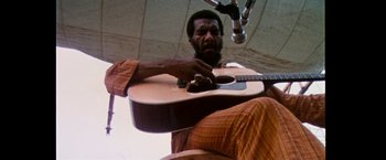 Movie still from “Woodstock” (1970), directed by Michael Wadleigh – A man holding a guitar while sitting on a chair; Medium shot, Low angle