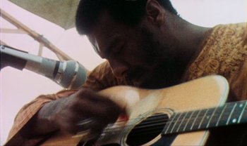 Movie still from “Woodstock” (1970), directed by Michael Wadleigh – A man playing a guitar while holding a microphone; Extreme Close Up shot, Low angle