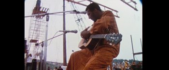 Movie still from “Woodstock” (1970), directed by Michael Wadleigh – A man playing a guitar on a stage with microphones in front of him; Medium shot, Low angle