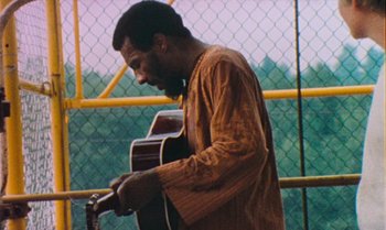 Movie still from “Woodstock” (1970), directed by Michael Wadleigh – A man playing an acoustic guitar in front of a chain link fence; Medium shot, Low angle