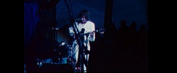 Movie still from “Woodstock” (1970), directed by Michael Wadleigh – A man in a white shirt is playing a guitar; Medium shot, Low angle