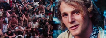 Movie still from “Woodstock” (1970), directed by Michael Wadleigh – A group of people sitting on the ground and a person standing in front of a group of other people; Close Up shot, High angle