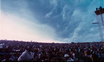 Movie still from “Woodstock” (1970), directed by Michael Wadleigh – A crowd of people sitting in a field under a cloudy sky; Extreme Wide shot, High angle