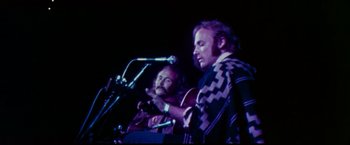 Movie still from “Woodstock” (1970), directed by Michael Wadleigh – Two men sitting at a microphone on a stage with microphones; Medium shot, Low angle