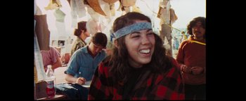 Movie still from “Woodstock” (1970), directed by Michael Wadleigh – A woman with a headband smiles in front of other people; Medium shot, Over the shoulder angle