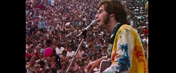 Movie still from “Woodstock” (1970), directed by Michael Wadleigh – A man is singing into a microphone in front of a large crowd; Medium shot, High angle
