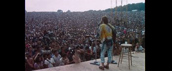 Movie still from “Woodstock” (1970), directed by Michael Wadleigh – A crowd of people sitting and standing in front of a microphone; Wide shot, High angle