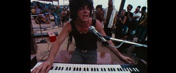 Movie still from “Woodstock” (1970), directed by Michael Wadleigh – A person sitting at a keyboard with a microphone in front of them; Medium shot, Low angle