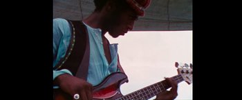 Movie still from “Woodstock” (1970), directed by Michael Wadleigh – A man playing a bass guitar on a sunny day; Medium shot, Low angle