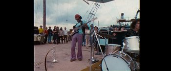Movie still from “Woodstock” (1970), directed by Michael Wadleigh – A man playing a bass guitar on a stage; Wide shot, Low angle