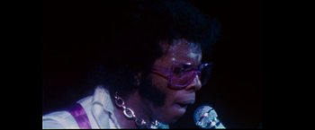 Movie still from “Woodstock” (1970), directed by Michael Wadleigh – A person with a microphone wearing glasses and a chain around their neck; Close Up shot, Low angle