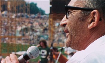 Movie still from “Woodstock” (1970), directed by Michael Wadleigh – An older man is speaking into a microphone at an outdoor concert; Close Up shot, Over the shoulder angle
