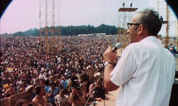 Movie still from “Woodstock” (1970), directed by Michael Wadleigh – A man is speaking to a large crowd of people; Wide shot, Over the shoulder angle